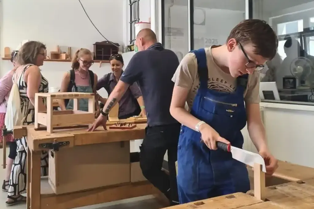 Azubi arbeitet in Holzwerkstatt, im Hintergrund Zeigt Meister Besuchern Werkbank