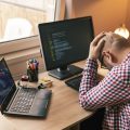 Software developer sitting at his desk while working in home office, holding head in hands, stressed out because of failure and code malfunction